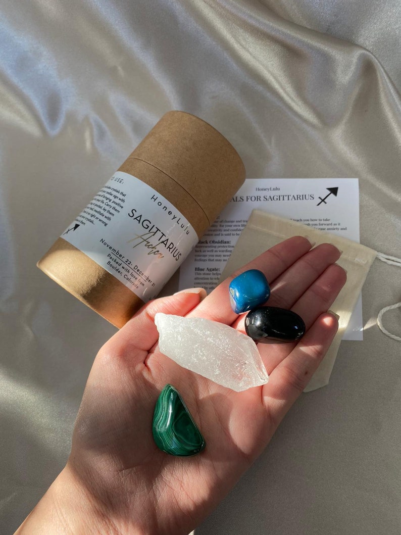 May include: A set of four crystals for the Sagittarius zodiac sign. The crystals are blue agate, black obsidian, clear quartz, and malachite. The crystals are arranged in a hand, with a small brown paper bag and a card with the text "Crystals for Sagittarius" in the background.