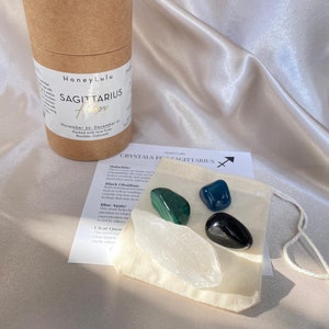 May include: A set of crystals for Sagittarius, including malachite, black obsidian, blue agate, and clear quartz. The crystals are in a small white pouch with a tag that reads "Crystals for Sagittarius".