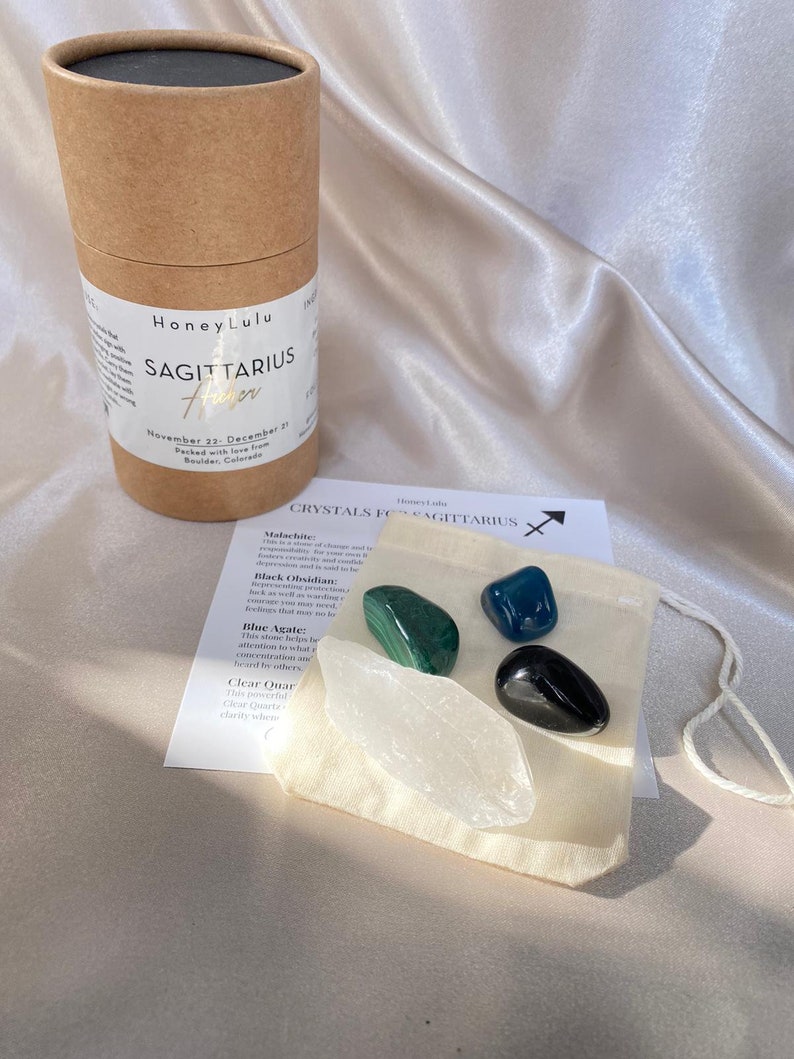 May include: A set of crystals for Sagittarius, including malachite, black obsidian, blue agate, and clear quartz. The crystals are in a small white pouch with a tag that reads "Crystals for Sagittarius".