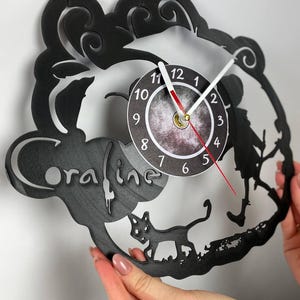 Puede incluir: Reloj de vinilo negro con una silueta de una ni&ntilde;a con un gato caminando frente a una luna. La esfera del reloj es blanca con n&uacute;meros y manecillas negros. El disco tiene la palabra "oraline" escrita en &eacute;l.