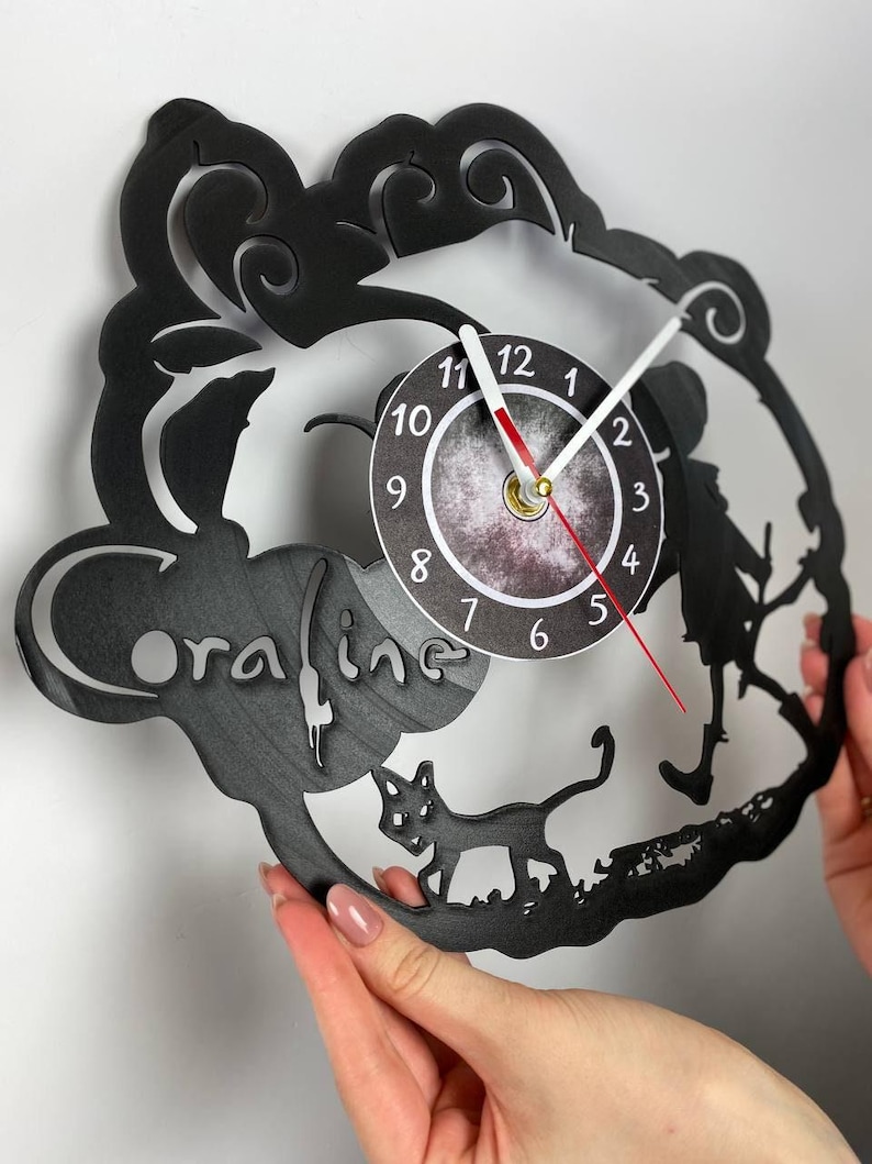 Puede incluir: Reloj de vinilo negro con una silueta de una ni&ntilde;a con un gato caminando frente a una luna. La esfera del reloj es blanca con n&uacute;meros y manecillas negros. El disco tiene la palabra "oraline" escrita en &eacute;l.