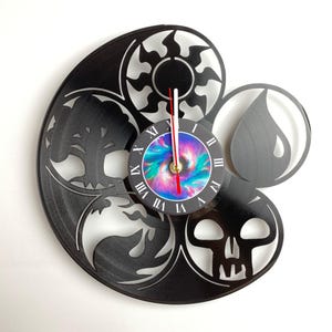 May include: A black vinyl record clock featuring the Magic: The Gathering symbols for the five colors of mana: white, blue, black, red, and green. The clock face is a swirling galaxy design with Roman numerals.