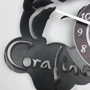 Puede incluir: Reloj de vinilo negro con una silueta de un gato y la palabra "oraline" recortada. La esfera del reloj es visible en el fondo.