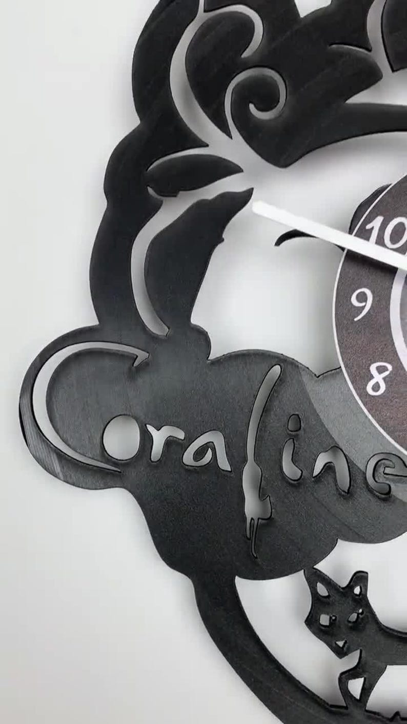 Puede incluir: Reloj de vinilo negro con una silueta de un gato y la palabra "oraline" recortada. La esfera del reloj es visible en el fondo.