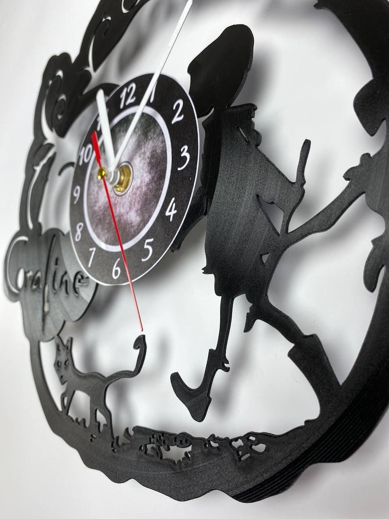 Puede incluir: Reloj de vinilo negro con una silueta de una ni&ntilde;a caminando con un gato. La esfera del reloj tiene un fondo negro con n&uacute;meros y manecillas blancas.