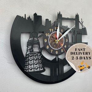 May include: Black vinyl record clock featuring a silhouette of the London skyline, including the Tower Bridge, and a Dalek from the Doctor Who television series. The clock face is a steampunk-style design with Roman numerals and a gear-shaped center. The clock is surrounded by a black frame with a white sticker that reads "FAST DELIVERY 2-3 DAYS".