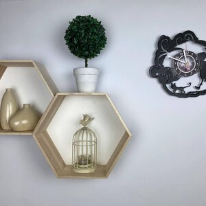 Puede incluir: Dos estantes hexagonales de madera con interiores blancos, montados en una pared blanca. Los estantes est&aacute;n decorados con jarrones y una jaula para p&aacute;jaros. Un reloj de vinilo negro con una esfera blanca y agujas negras est&aacute; montado en la pared.