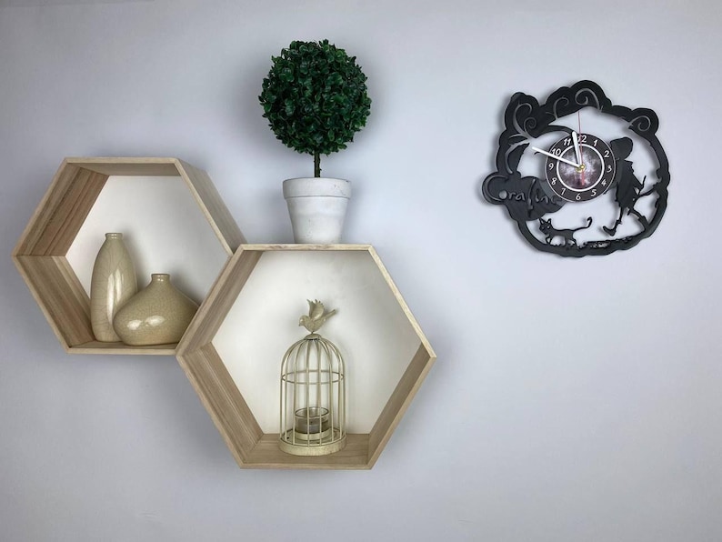 Puede incluir: Dos estantes hexagonales de madera con interiores blancos, montados en una pared blanca. Los estantes est&aacute;n decorados con jarrones y una jaula para p&aacute;jaros. Un reloj de vinilo negro con una esfera blanca y agujas negras est&aacute; montado en la pared.