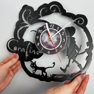 Puede incluir: Reloj de vinilo negro con una silueta de una ni&ntilde;a caminando con un gato. La esfera del reloj es un c&iacute;rculo blanco con n&uacute;meros negros y manecillas. El reloj est&aacute; etiquetado como "oraline".
