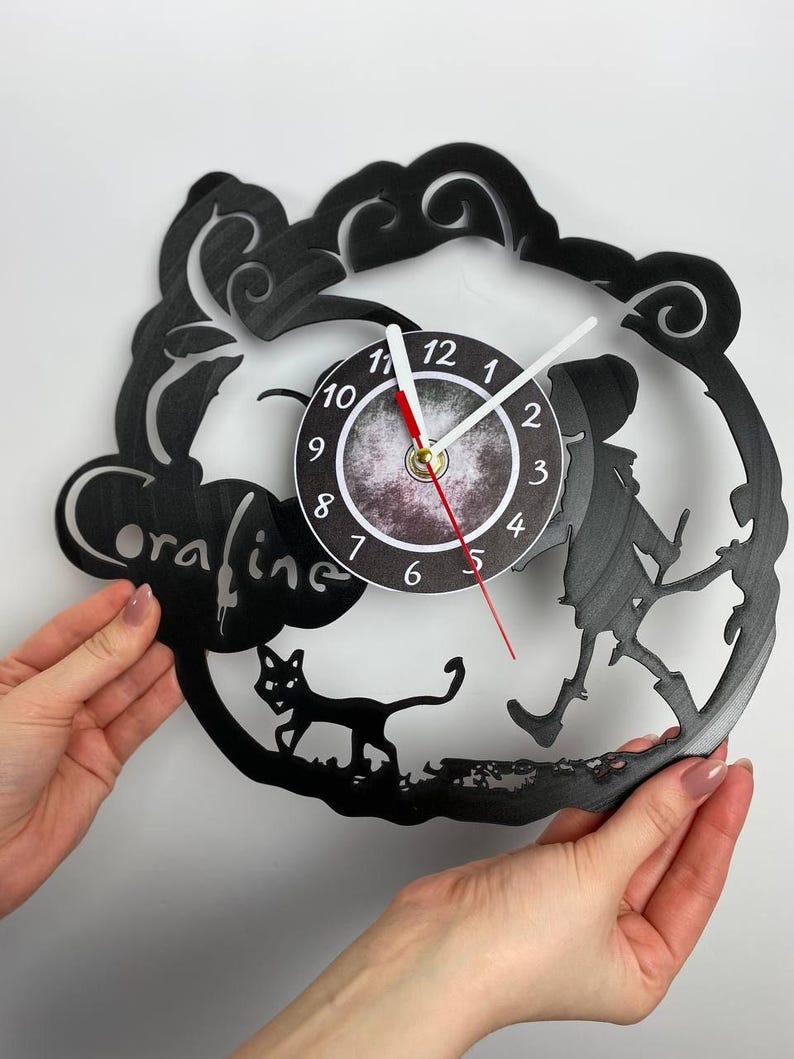 Puede incluir: Reloj de vinilo negro con una silueta de una ni&ntilde;a caminando con un gato. La esfera del reloj es un c&iacute;rculo blanco con n&uacute;meros negros y manecillas. El reloj est&aacute; etiquetado como "oraline".
