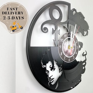 May include: A black vinyl record clock featuring a portrait of Prince with his iconic symbol. The clock face is white with black numbers and hands.