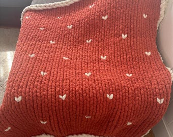 May include: A chunky knit blanket in a warm, reddish-orange hue, adorned with small white heart-shaped accents. The blanket has a thick, textured appearance and is edged with a cream-colored border, creating a cozy, handmade aesthetic.