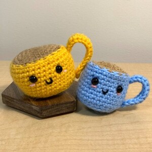 Puede incluir: Dos tazas de café de ganchillo con caras sonrientes. Una es amarilla, la otra azul. Ambas tienen 'café' marrón en el interior y asas amarillas o azules. Las tazas están sobre un pequeño bloque de madera.