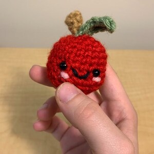 May include: A red crocheted apple with a green leaf and a smiling face. The apple has black eyes and pink cheeks.