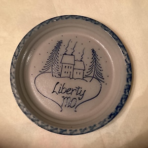 May include: A round, ceramic plate with a blue rim and a hand-drawn design. The design features two houses, trees, and the words "Liberty mo" in a heart shape, all in blue. The plate has a light gray background.
