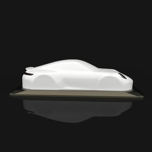 May include: A white, smooth, abstract car sculpture featuring a sleek, aerodynamic silhouette with a rounded roofline, a small rear spoiler, and a single visible circular headlight, mounted on a dark, rectangular base against a black background.