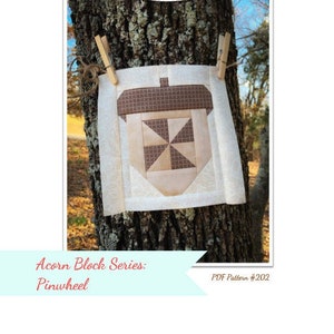May include: A quilt block pattern for a pinwheel design, part of the Acorn Block Series. The block is made of brown, beige, and white fabric and is hanging from a tree branch. The text "Acorn Block Series: Pinwheel" and "PDF Pattern #202" is visible.