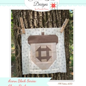 Op de afbeelding: Een quiltblokpatroon voor een churn dash-ontwerp, onderdeel van de Acorn Block Series. Het blok is gemaakt van bruine, witte en beige stof en wordt getoond op een boomstam. De tekst "PDF Pattern #212" is zichtbaar in de rechterbenedenhoek.
