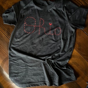 Op de afbeelding: Een donkergrijs T-shirt met een rood ontwerp van een kip en het woord "Ohio" gespeld in kleine voetafdrukken.
