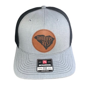 May include: Gray and black trucker hat with a brown leather patch featuring the text "Travelers Rest" and an outline of the state of South Carolina. The hat is adjustable and has the Richardson 112 logo.