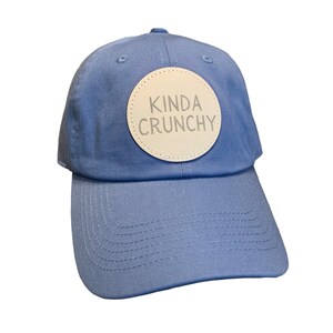 May include: A light blue baseball cap with a white circle patch that says "Kinda Crunchy" in gray lettering.