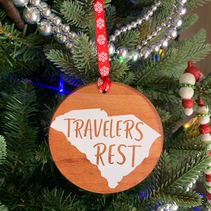 May include: A circular wooden Christmas decoration featuring the words "Travelers Rest" in white, and a white outline of South Carolina. It hangs from a red ribbon with white snowflakes, on a Christmas tree with silver baubles.