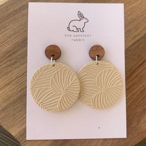 May include: A pair of cream-colored, round earrings with a leaf pattern. The earrings are made of polymer clay and have wooden tops.