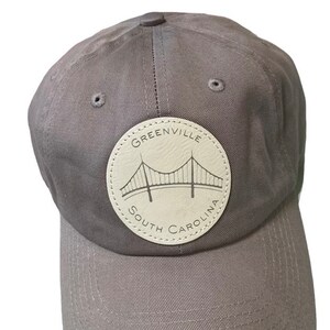 May include: A gray baseball cap with a beige leather patch featuring the text "Greenville South Carolina" and a stylized bridge design.