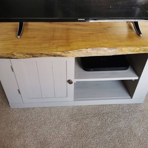May include: A wooden TV stand with a natural edge top. The top is a light brown color, and the base is painted a light gray. The stand has a cabinet door on the left and an open shelf on the right. A black TV sits on top.
