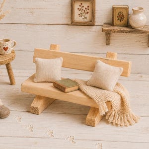 Può includere: Una panchina in legno in miniatura con due cuscini in tessuto beige, un piccolo libro verde e una coperta beige con frange. Accanto alla panchina ci sono due scarpine in maglia marrone, uno sgabello rotondo in legno, un vaso e una tazza a cuori.