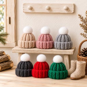 Könnte beinhalten: Eine Sammlung von sechs kleinen, grob gestrickten Mützen, die jeweils mit einem flauschigen weißen Bommel versehen sind. Die Mützen sind in zwei Reihen angeordnet und in verschiedenen Unifarben wie Beige, Altrosa, Grau, Anthrazit, Rot und Grün.
