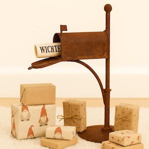 Puede incluir: Un buzón de metal marrón oxidado con un pergamino "WICHTE" está rodeado de regalos envueltos. Algunos regalos están envueltos en papel marrón liso, otros con un patrón de gnomos. La escena se desarrolla sobre una superficie blanca y esponjosa.