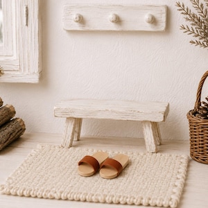 May include: A miniature wooden bench with a distressed white finish, a pair of small brown slide sandals, and a rectangular cream-colored woven rug. The scene includes a wall-mounted coat rack with three knobs and a small brown wicker basket.