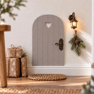 May include: A small arched grey wooden door with a heart-shaped cutout, a brass handle, and a keyhole. Beside the door is a wall-mounted lantern, a small bundle of pine branches with a pinecone, and two brown gift boxes with white tree patterns.