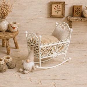 Könnte beinhalten: Eine weiße Miniaturwiege aus Metall mit Gitterdesign, die eine beigefarbene Kunstfelldecke und ein kleines Kissen enthält. Eine beige Zwergenfigur sitzt neben der Wiege und ein Paar kleine, beigefarbene Strickschühchen stehen auf dem Boden.