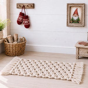 Peut inclure: Un tapis rectangulaire en macramé blanc cassé présentant un motif de nœuds répétitif et des bords à franges. Le tapis est posé sur un sol clair dans une pièce décorée d'un panier en osier rempli de bûches, de moufles rouges et d'un cadre.