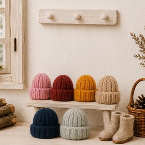 Könnte beinhalten: Eine Sammlung von sechs kleinen, gestrickten Mützen in Rosa, Dunkelrot, Senfgelb, Beige, Marineblau und Hellgrau, begleitet von einem Paar beiger Filzstiefel. Jede Mütze zeichnet sich durch eine gerippte Textur und ein umgeschlagenes Bündchen aus.