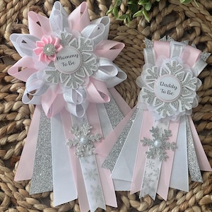 Könnte beinhalten: Zwei Babyparty-Rosetten mit rosa, weißen und silbernen Bändern. Eine Rosette trägt die Aufschrift "Mommy To Be", die andere "Daddy To Be". Jede Rosette hat ein Schneeflocken-Design und einen Perlenakzent.