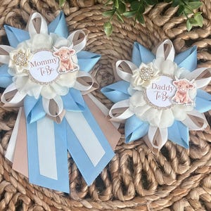 Könnte beinhalten: Zwei dekorative Anstecknadeln aus Ripsband in Hellblau, Weiß und Beige. Jede Anstecknadel hat ein weißes, rundes Emblem mit glitzerndem Goldrand, eine kleine Stoffblume mit Strassstein und eine Highland-Rind-Illustration. Eine Anstecknadel trägt die Aufschrift Mommy To Be, die andere Daddy To Be.