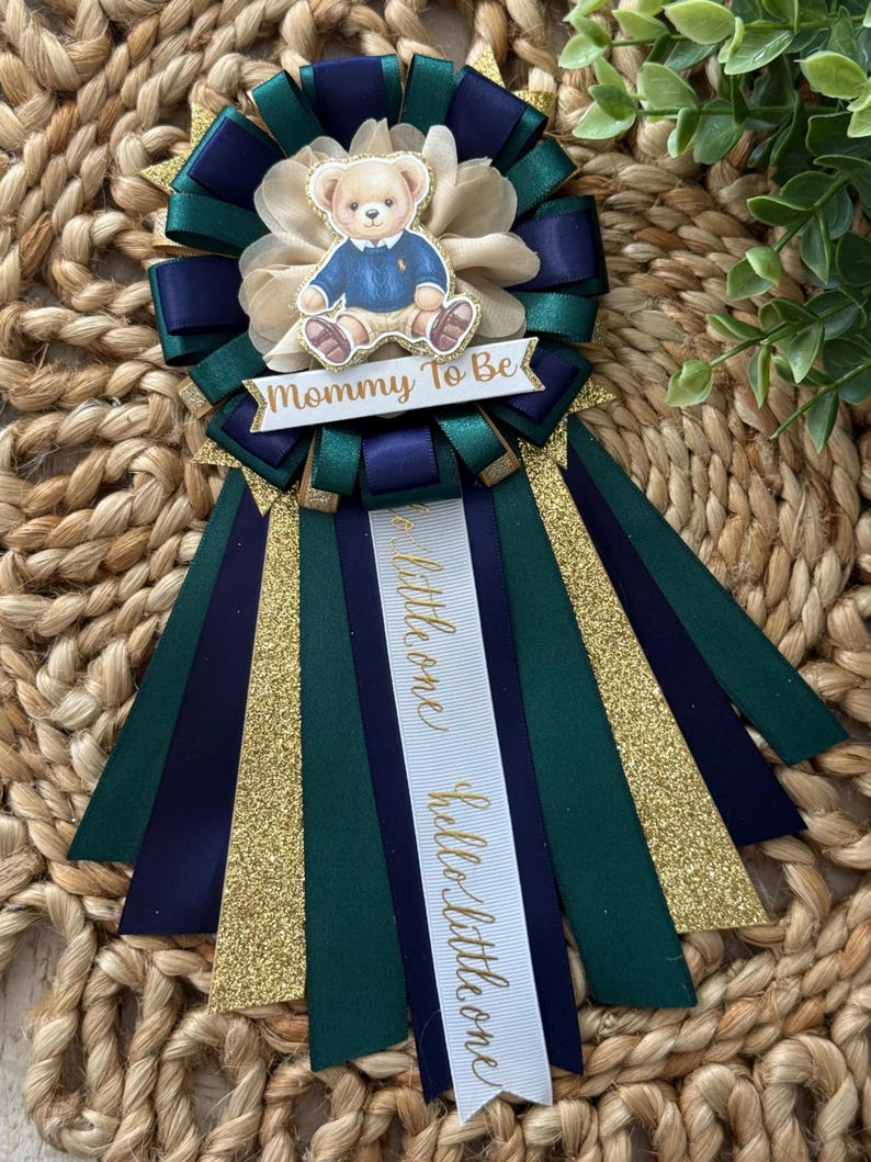 K&ouml;nnte beinhalten: Ein Babyparty-Band mit einem Teddyb&auml;ren in einem blauen Pullover. Das Band hat marineblaue, smaragdgr&uuml;ne und goldglitzernde Akzente. Die Texte "Mommy To Be" und "hello little one" sind auf dem Band zu sehen.