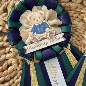 K&ouml;nnte beinhalten: Babyparty-Rosette mit Teddyb&auml;r-Illustration. Die Rosette hat marineblaue, smaragdgr&uuml;ne und goldglitzernde B&auml;nder. Der Text "Mommy To Be" ist auf einem wei&szlig;en Banner zu sehen. Ein zweites Band sagt "a little one".