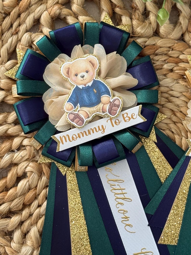 K&ouml;nnte beinhalten: Babyparty-Rosette mit Teddyb&auml;r-Illustration. Die Rosette hat marineblaue, smaragdgr&uuml;ne und goldglitzernde B&auml;nder. Der Text "Mommy To Be" ist auf einem wei&szlig;en Banner zu sehen. Ein zweites Band sagt "a little one".