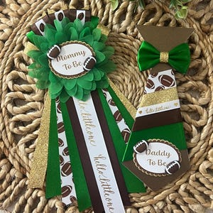 Könnte beinhalten: Ein grün-brauner Ansteck-Button mit der Aufschrift Mommy To Be und ein krawattenförmiger Button mit der Aufschrift Daddy To Be. Beide zeigen Football-Grafiken, goldene Glitzerakzente und Bänder mit dem Schriftzug hello little one.