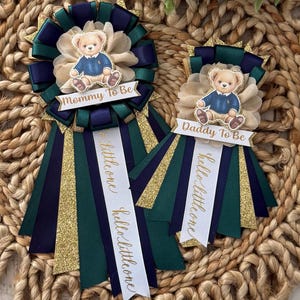 K&ouml;nnte beinhalten: Zwei Babyparty-Rosetten mit Teddyb&auml;r-Akzenten. Auf einer Rosette steht "Mommy To Be" und auf der anderen "Daddy To Be". Beide haben marineblaue, gr&uuml;ne und goldglitzernde B&auml;nder. Auf den B&auml;ndern steht auch "hello little one".