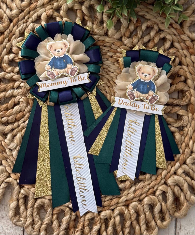 K&ouml;nnte beinhalten: Zwei Babyparty-Rosetten mit Teddyb&auml;r-Akzenten. Auf einer Rosette steht "Mommy To Be" und auf der anderen "Daddy To Be". Beide haben marineblaue, gr&uuml;ne und goldglitzernde B&auml;nder. Auf den B&auml;ndern steht auch "hello little one".