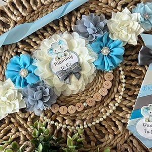 Könnte beinhalten: Ein blaues und weißes Babyparty-Band mit den Worten "Mommy To Be" und einem kleinen Elefanten-Design. Eine passende Krawatte mit den Worten "Daddy To Be" und einem kleinen Elefanten-Design. Eine Perlenkette mit Holzbuchstabenperlen, die "Liam Mateo" buchstabieren.