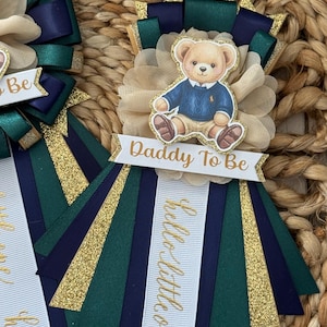 K&ouml;nnte beinhalten: Babyparty-Rosette mit einer Teddyb&auml;r-Illustration in einem blauen Pullover. Die Rosette hat marineblaue, gr&uuml;ne und goldene B&auml;nder mit dem Text "Daddy To Be" und "hello little one" in goldener Schrift. Der Hintergrund hat eine gewebte Textur.