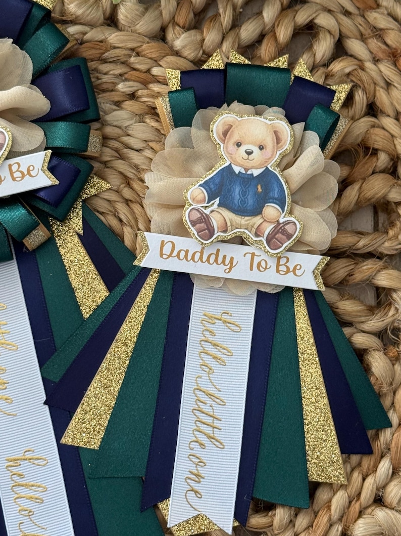 K&ouml;nnte beinhalten: Babyparty-Rosette mit einer Teddyb&auml;r-Illustration in einem blauen Pullover. Die Rosette hat marineblaue, gr&uuml;ne und goldene B&auml;nder mit dem Text "Daddy To Be" und "hello little one" in goldener Schrift. Der Hintergrund hat eine gewebte Textur.