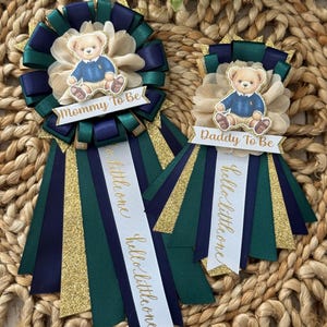 K&ouml;nnte beinhalten: Zwei Babyparty-Ansteckblumen mit Teddyb&auml;r-Akzenten. Eine Ansteckblume tr&auml;gt die Aufschrift "Mommy To Be", die andere "Daddy To Be". Die Ansteckblumen sind marineblau, gr&uuml;n, gold und cremefarben mit B&auml;ndern mit der Aufschrift "hello little one".