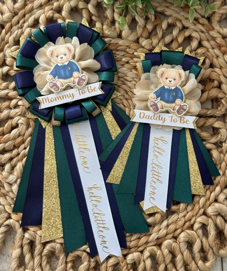 K&ouml;nnte beinhalten: Zwei Babyparty-Ansteckblumen mit Teddyb&auml;r-Akzenten. Eine Ansteckblume tr&auml;gt die Aufschrift "Mommy To Be", die andere "Daddy To Be". Die Ansteckblumen sind marineblau, gr&uuml;n, gold und cremefarben mit B&auml;ndern mit der Aufschrift "hello little one".
